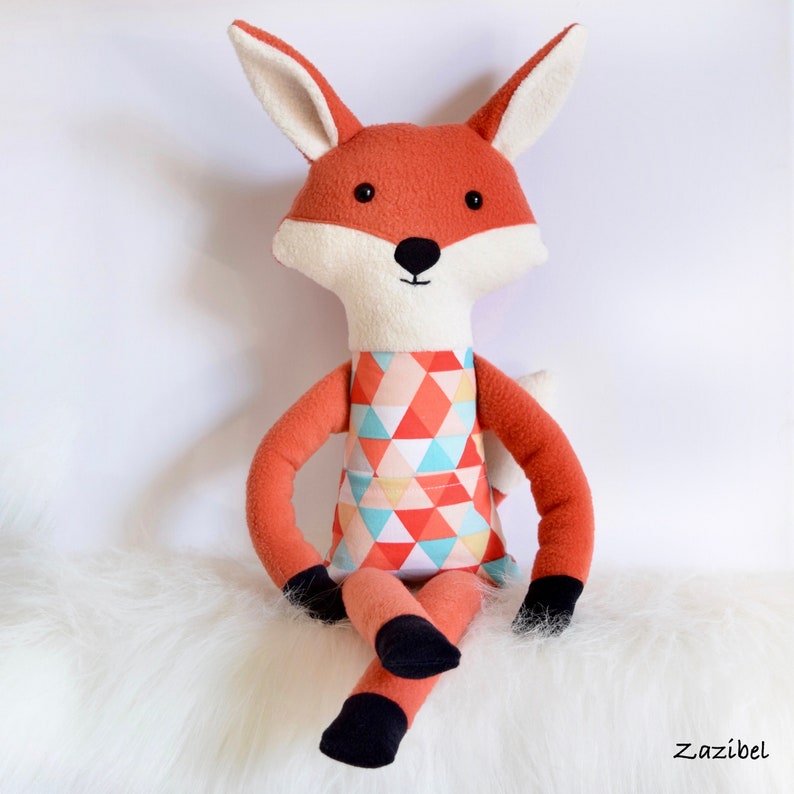May include: A plush fox toy with orange fur, black eyes, and a white face. The fox is wearing a colorful geometric patterned dress and is sitting on a white fluffy surface.