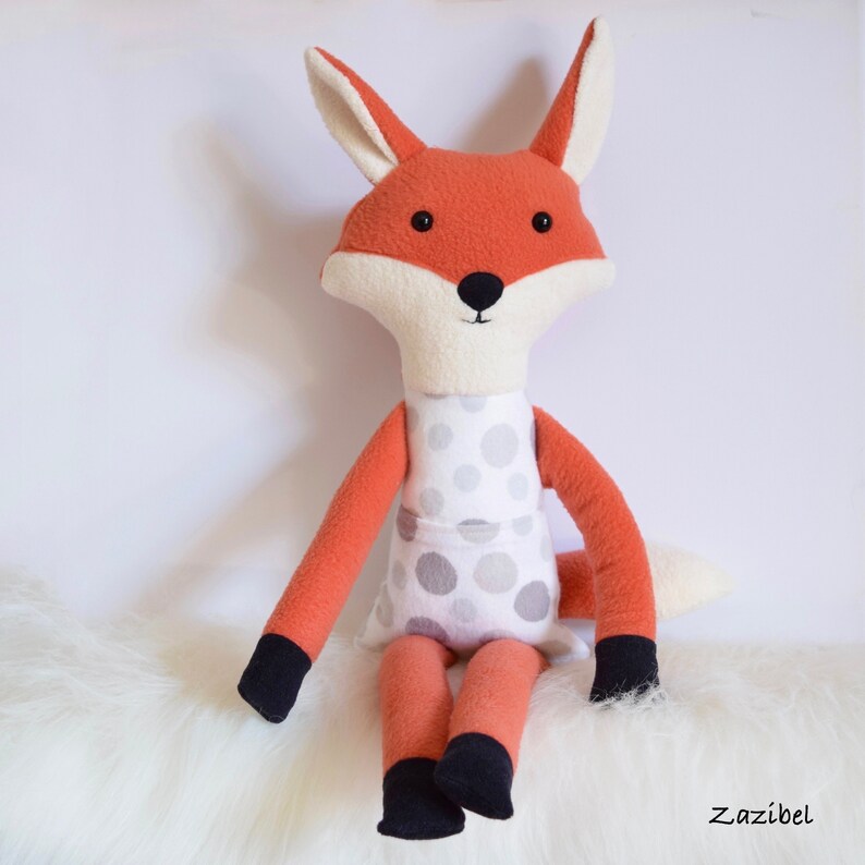 May include: A plush fox toy with orange fur, white face, and black eyes. The fox is wearing a white dress with gray polka dots and has black feet.