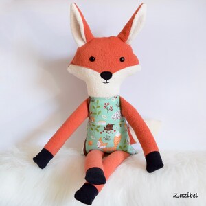 May include: A plush fox toy with orange fur, white face, and black eyes. The fox is wearing a green and white dress with a woodland animal print. The toy is sitting on a white fluffy surface.