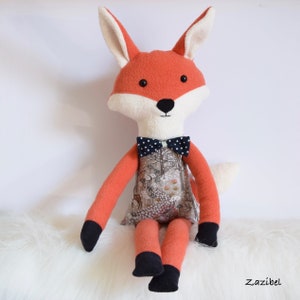 May include: A plush fox toy with orange fur, white facial features, and a black and white bow tie. The fox is wearing a dress with a woodland print and is sitting on a white fluffy surface.