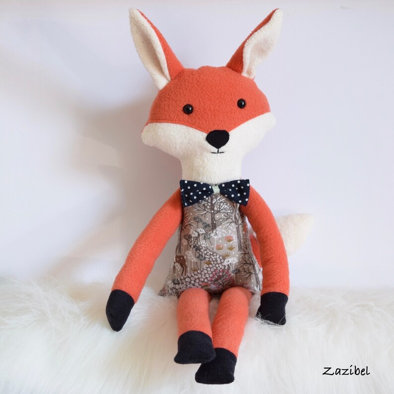 May include: A plush fox toy with orange fur, white facial features, and a black and white bow tie. The fox is wearing a dress with a woodland print and is sitting on a white fluffy surface.