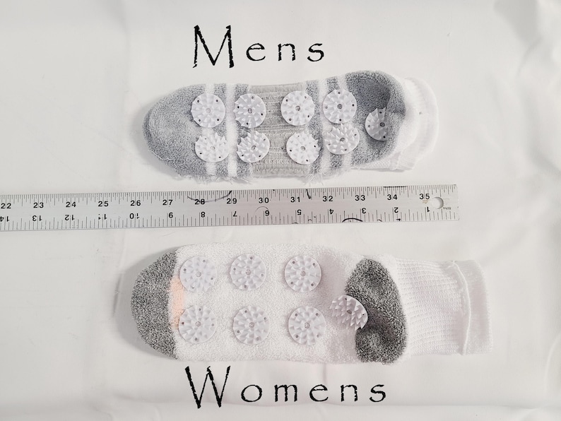 Pu&ograve; includere: Due calzini con dischi di agopressione in plastica bianca sulle suole, etichettati Mens e Womens. Un righello &egrave; posizionato tra loro, mostrando una lunghezza di circa 33 cm per il calzino superiore e 28 cm per quello inferiore.