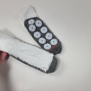 Pu&ograve; includere: Un paio di calzini bianchi e grigi con polsino a coste bianco e suola grigia. La parte inferiore di un calzino mostra otto dischi circolari per agopressione in plastica bianca con piccole punte e un piccolo dettaglio ricamato in rosso.