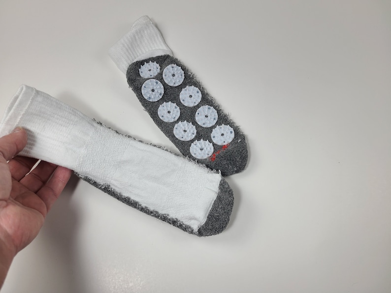 Pu&ograve; includere: Un paio di calzini bianchi e grigi con polsino a coste bianco e suola grigia. La parte inferiore di un calzino mostra otto dischi circolari per agopressione in plastica bianca con piccole punte e un piccolo dettaglio ricamato in rosso.