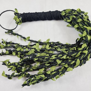 May include: A black handle wrapped in cord with a circular loop at the end, featuring multiple long, braided black strands adorned with small, vibrant green fabric leaves that extend from the handle to create a vine-like appearance.