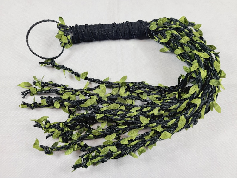 May include: A black handle wrapped in cord with a circular loop at the end, featuring multiple long, braided black strands adorned with small, vibrant green fabric leaves that extend from the handle to create a vine-like appearance.