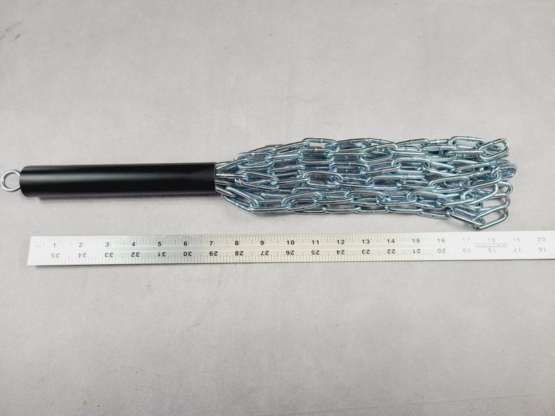 Puede incluir: Un mango de metal negro con un mayal de cadena plateada. La cadena est&aacute; hecha de eslabones de metal entrelazados. Se coloca una regla junto al mayal para mostrar la longitud del objeto.