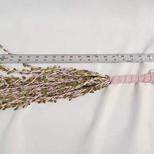 May include: A multi-strand whip featuring light pink cord intertwined with small green fabric leaves. The handle is wrapped in matching light pink material with a loop at the end. A metal ruler placed above shows the object is 18 inches long.