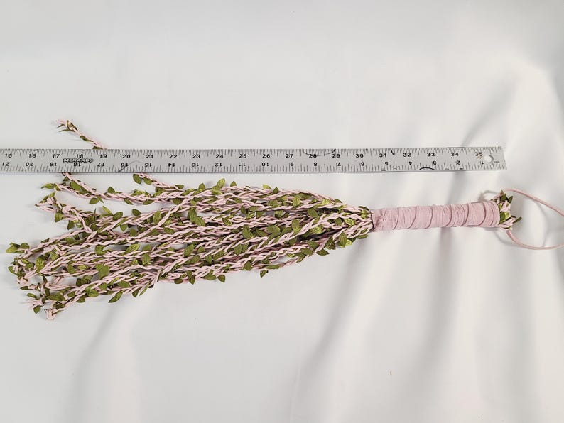 May include: A multi-strand whip featuring light pink cord intertwined with small green fabric leaves. The handle is wrapped in matching light pink material with a loop at the end. A metal ruler placed above shows the object is 18 inches long.