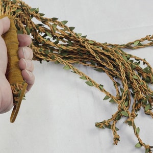 May include: A multi-strand whip featuring a tan suede-wrapped handle and numerous long, braided tan cords adorned with small, pointed green fabric leaves. The strands extend from the base, creating a vine-like appearance with textured foliage.