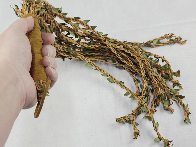 May include: A multi-strand whip featuring a tan suede-wrapped handle and numerous long, braided tan cords adorned with small, pointed green fabric leaves. The strands extend from the base, creating a vine-like appearance with textured foliage.