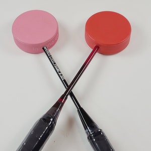 May include: Two badminton shuttlecocks with red and pink tops are crossed. The shuttlecocks have black handles and the text "TEMPERED STEEL FRAME C-305" is visible on one. The tops are round and have a textured edge.