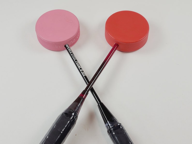 May include: Two badminton shuttlecocks with red and pink tops are crossed. The shuttlecocks have black handles and the text "TEMPERED STEEL FRAME C-305" is visible on one. The tops are round and have a textured edge.