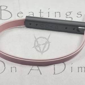 Może przedstawiać: Różowy pasek skórzany z czarną plastikową klamrą. Klamra ma wytłoczone słowo "ROAD".