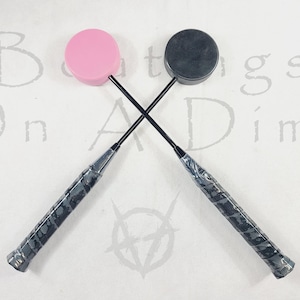 May include: Two badminton rackets crossed, one with a pink head and the other with a dark grey head. The handles are wrapped in a grey grip. The background has the words "Beatings On A Dime" in a faded font.