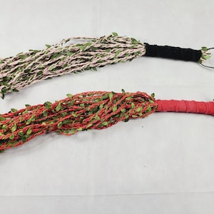 May include: Two floggers featuring multiple strands of light pink and red cord intertwined with small green artificial leaves. Each flogger has a wrapped handle, one in black and one in red, with a matching loop at the end for hanging.