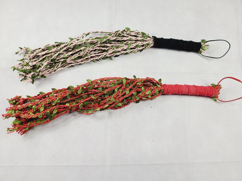 May include: Two floggers featuring multiple strands of light pink and red cord intertwined with small green artificial leaves. Each flogger has a wrapped handle, one in black and one in red, with a matching loop at the end for hanging.