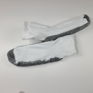 Pu&ograve; includere: Un paio di calzini alla caviglia con una sezione superiore bianca e una suola testurizzata grigio scuro. I calzini presentano un bordo a coste sulla parte superiore e un design bicolore con il tessuto grigio sulla parte inferiore e sulla punta.