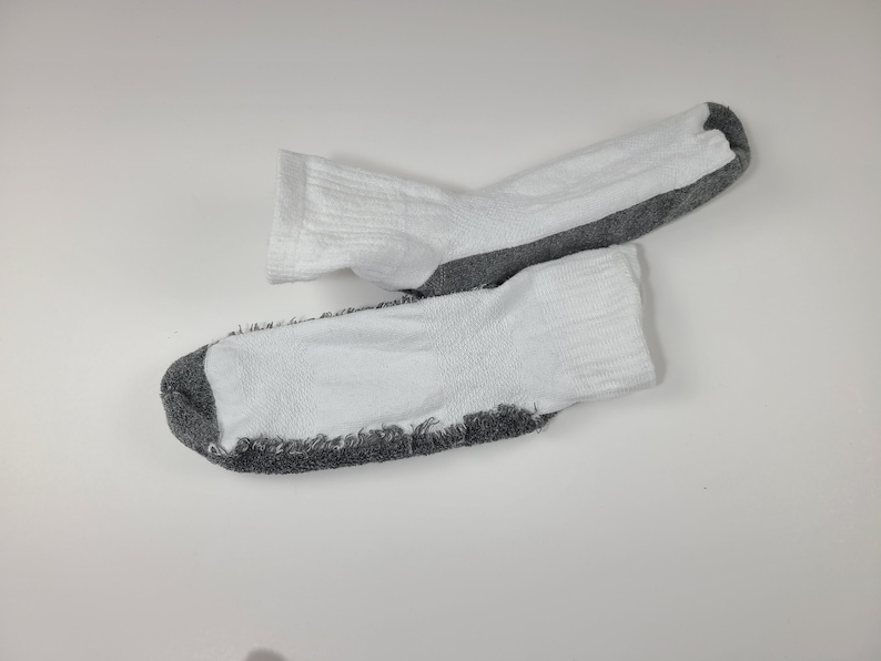 Pu&ograve; includere: Un paio di calzini alla caviglia con una sezione superiore bianca e una suola testurizzata grigio scuro. I calzini presentano un bordo a coste sulla parte superiore e un design bicolore con il tessuto grigio sulla parte inferiore e sulla punta.