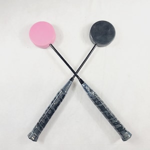 May include: Two badminton rackets crossed, one with a pink head and the other with a black head. The handles are wrapped in a textured black grip. The rackets are set against a white background.