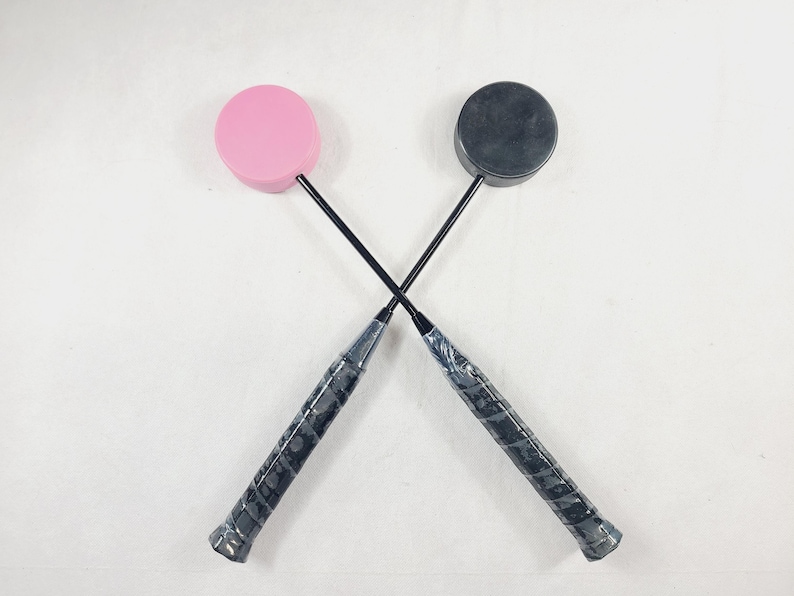 May include: Two badminton rackets crossed, one with a pink head and the other with a black head. The handles are wrapped in a textured black grip. The rackets are set against a white background.