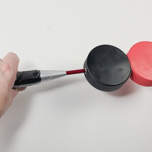 May include: A red metal rod attached to a black hockey puck, which is connected to a second red hockey puck. The rod features a silver-colored handle section wrapped in black tape, providing a grip for the connected circular pucks.
