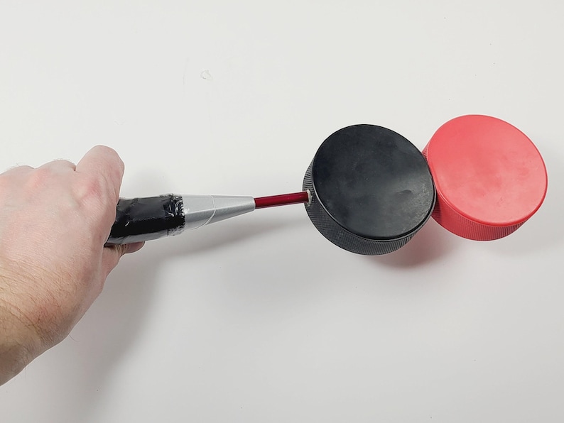 May include: A red metal rod attached to a black hockey puck, which is connected to a second red hockey puck. The rod features a silver-colored handle section wrapped in black tape, providing a grip for the connected circular pucks.
