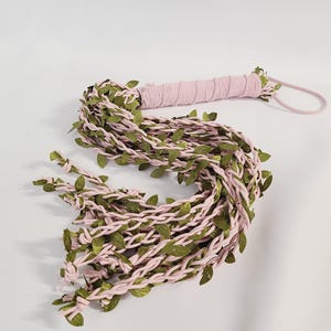 May include: A decorative whip featuring a handle wrapped in light pink fabric and a wrist loop. The main body consists of multiple braided light pink fabric strands intertwined with artificial green leaves, ending in knotted tips.