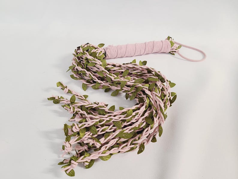 May include: A decorative whip featuring a handle wrapped in light pink fabric and a wrist loop. The main body consists of multiple braided light pink fabric strands intertwined with artificial green leaves, ending in knotted tips.
