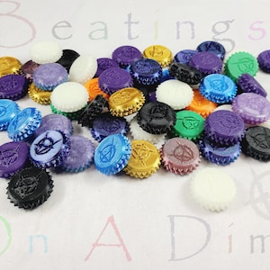 May include: A collection of colorful bottle caps in various shades of purple, blue, green, gold, orange, black, and white. Each cap features a unique embossed design. The caps are scattered on a white surface.