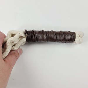 May include: A close-up of a brown leather cord wrapped around a white cotton cord. The cord is held in a hand.