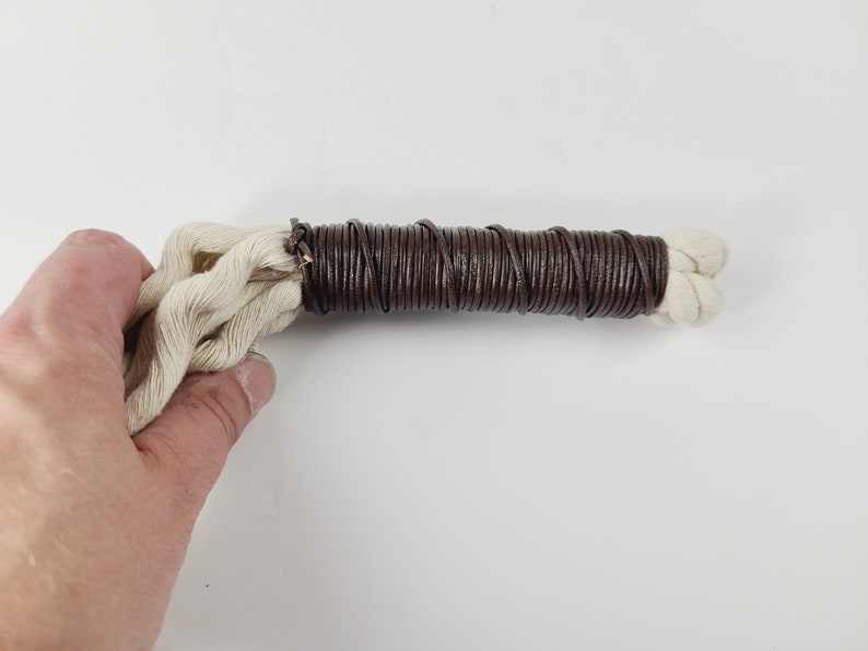 May include: A close-up of a brown leather cord wrapped around a white cotton cord. The cord is held in a hand.