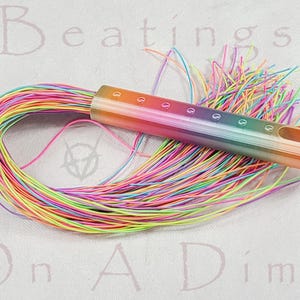 May include: A rainbow-colored bead loom needle with a bundle of colorful threads. The needle is a gradient of orange, yellow, green, blue, and purple. The text "Beatings On A Dime" is in the background.
