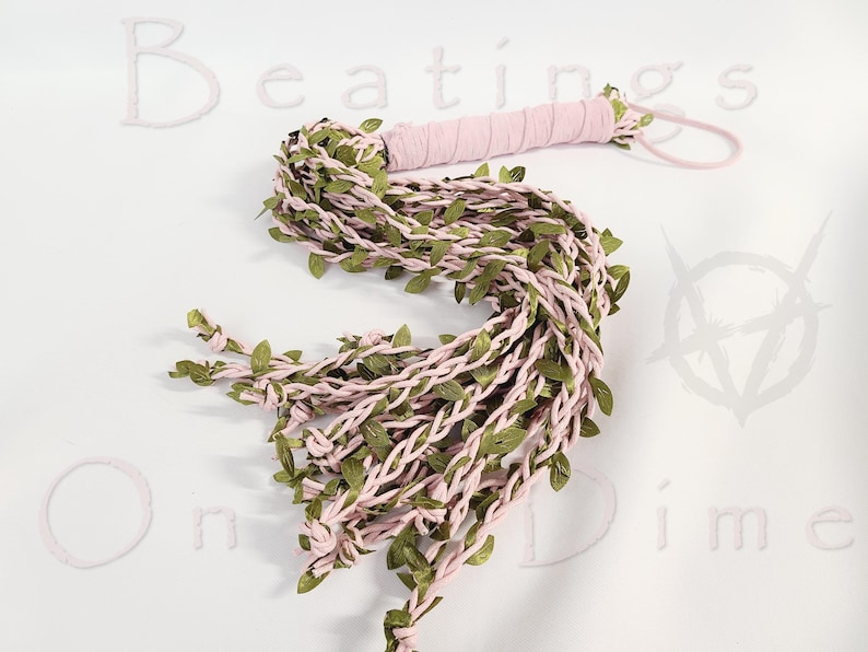 May include: A light pink braided whip featuring a wrapped handle and a wrist loop, adorned with numerous small green fabric leaves woven throughout the length of the strands. The text Beatings On a Dime is visible in the background.