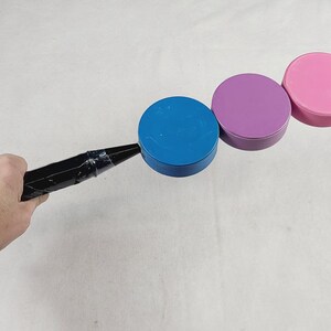 May include: A black handle attached to three circular plastic discs in blue, purple, and pink. The handle is secured with black tape to the blue disc, which is connected to the purple disc, followed by the pink disc in a linear arrangement.