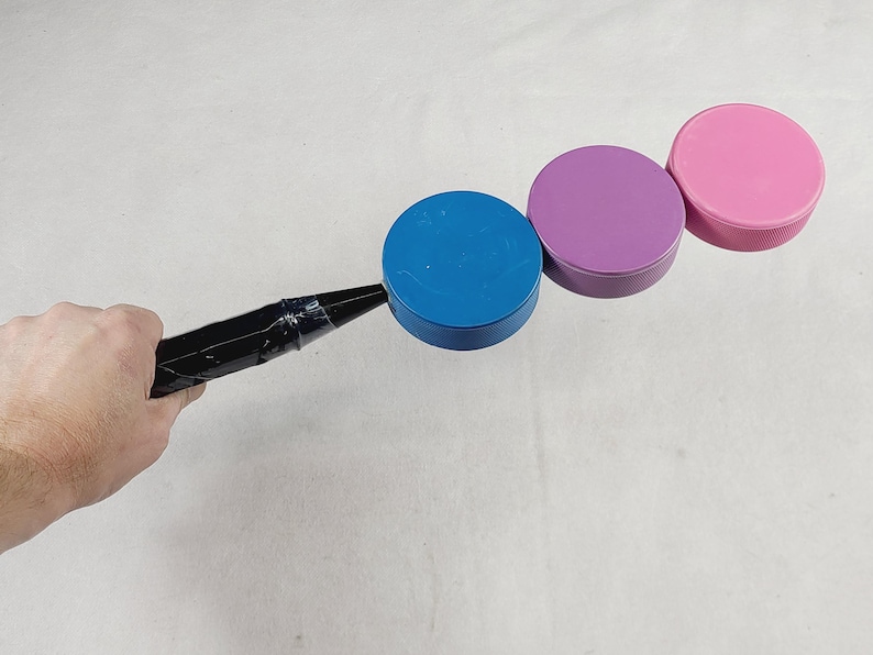 May include: A black handle attached to three circular plastic discs in blue, purple, and pink. The handle is secured with black tape to the blue disc, which is connected to the purple disc, followed by the pink disc in a linear arrangement.