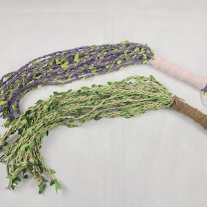 May include: Two multi-strand whips featuring braided cords adorned with small green fabric leaves. One whip has purple cords with a light pink wrapped handle, while the other has natural tan cords with a brown wrapped handle and a wrist loop.