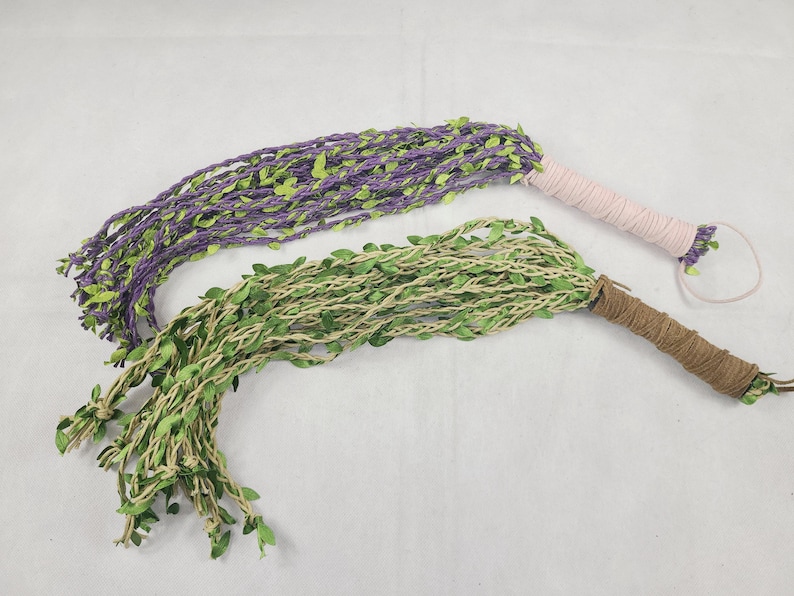 May include: Two multi-strand whips featuring braided cords adorned with small green fabric leaves. One whip has purple cords with a light pink wrapped handle, while the other has natural tan cords with a brown wrapped handle and a wrist loop.
