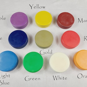 May include: Ten small, round, lidded containers in a variety of colours are displayed. The colours include purple, yellow, maroon, blue, gold, red, light blue, green, white, and orange. Each colour is labelled with handwritten text.