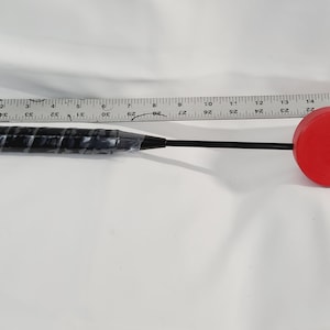 May include: A black and red mallet with a black handle and a red, circular striking head. The handle has a textured grip. A ruler is placed next to the mallet.