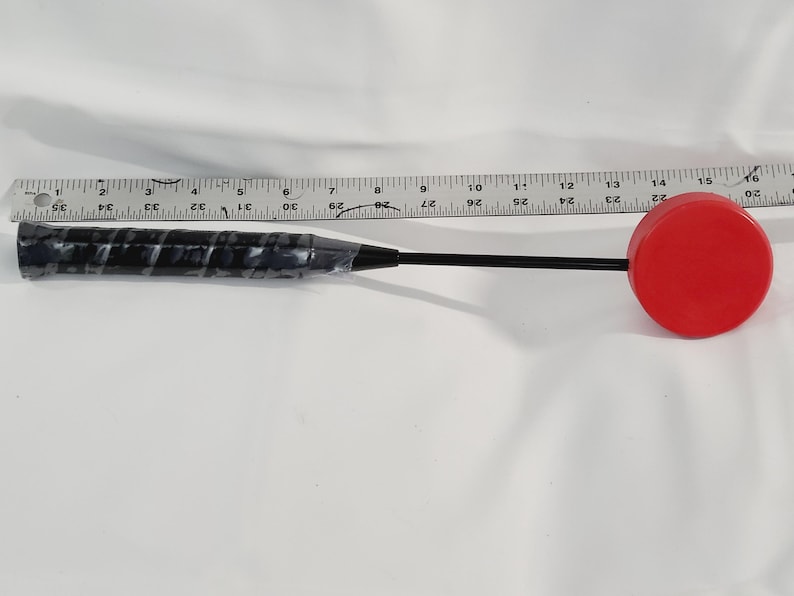 May include: A black and red mallet with a black handle and a red, circular striking head. The handle has a textured grip. A ruler is placed next to the mallet.