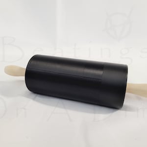 Peut inclure: Un rouleau à pâtisserie doté d'un cylindre noir mat et de poignées en bois clair à chaque extrémité. Le texte "Beatings On A Dime" est légèrement visible en arrière-plan derrière le rouleau à pâtisserie, qui est placé horizontalement.