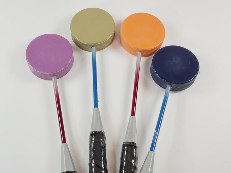 May include: Four bottle stoppers in various colours. Each stopper features a circular, solid-coloured top and a long, thin handle. The handles transition from a dark hue to a lighter shade. The stoppers have black grips.