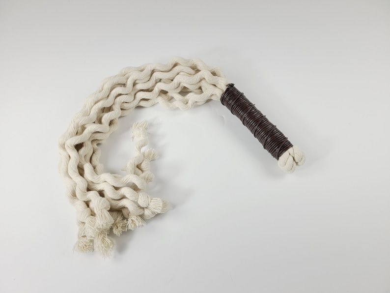 May include: A white cotton rope whip with a brown leather handle. The rope is braided and has a fringed end.
