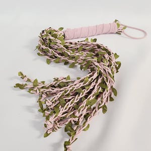 May include: A multi-strand whip featuring light pink fabric cords intertwined with small green artificial leaves. The handle is wrapped in matching light pink fabric and includes a thin pink loop at the end for holding or hanging the item.