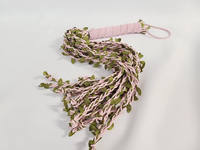 May include: A multi-strand whip featuring light pink fabric cords intertwined with small green artificial leaves. The handle is wrapped in matching light pink fabric and includes a thin pink loop at the end for holding or hanging the item.