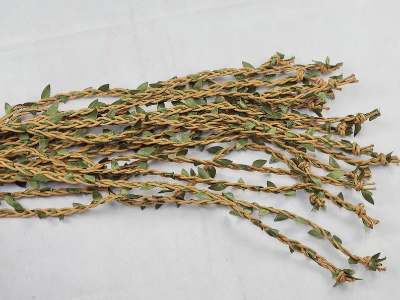 May include: A collection of multiple tan braided twine strands interwoven with small green fabric leaves. Each individual strand features a knotted end, creating a textured, vine-like appearance with a consistent brown and green color palette.