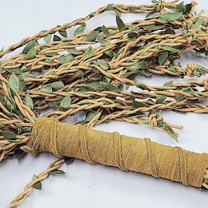 May include: A decorative bundle featuring multiple strands of tan braided cord intertwined with small green fabric leaves. The strands are gathered and secured at one end by a cylindrical handle wrapped in a textured tan suede-like material.