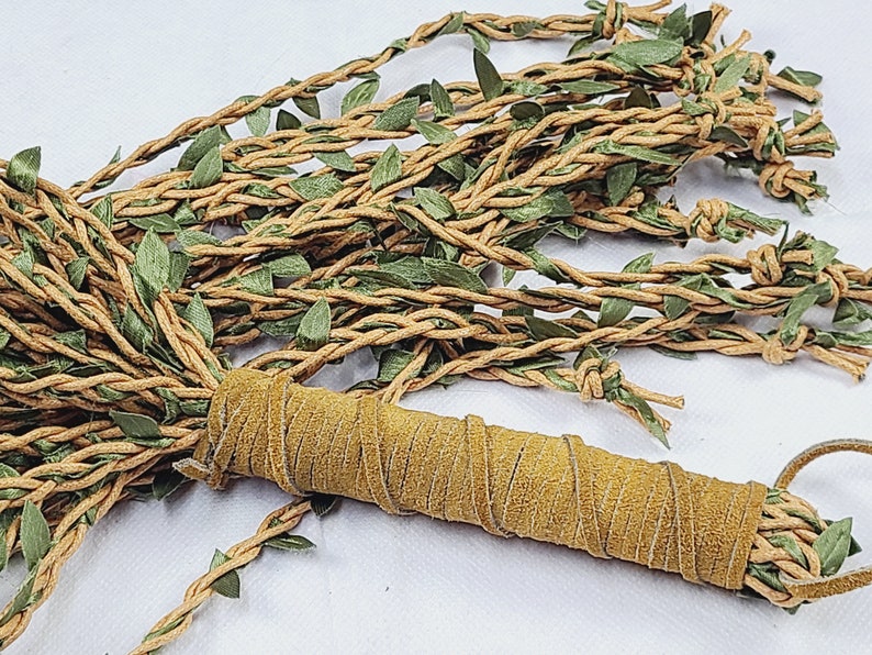 May include: A decorative bundle featuring multiple strands of tan braided cord intertwined with small green fabric leaves. The strands are gathered and secured at one end by a cylindrical handle wrapped in a textured tan suede-like material.