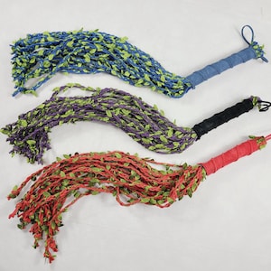 May include: Three decorative whips featuring long, braided strands in blue, purple, and red, each adorned with small green fabric leaves. The handles are wrapped in matching solid-colored fabric, and each whip includes a small loop at the end.